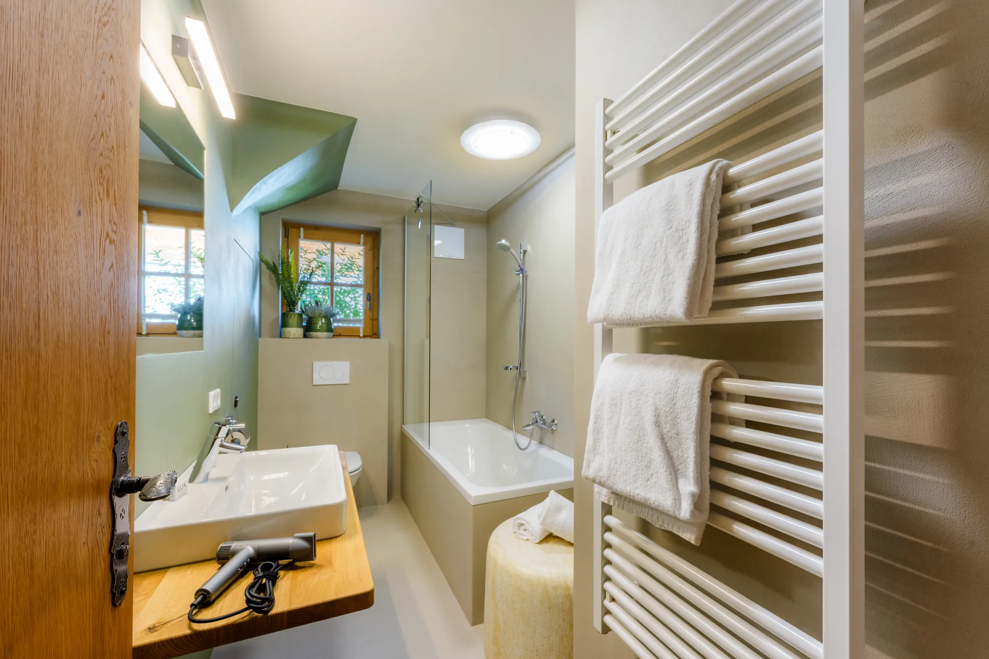 Helles Badezimmer mit Badewanne, Waschbecken auf Holzablage, Föhn und Handtuchheizkörper mit weißen Handtüchern