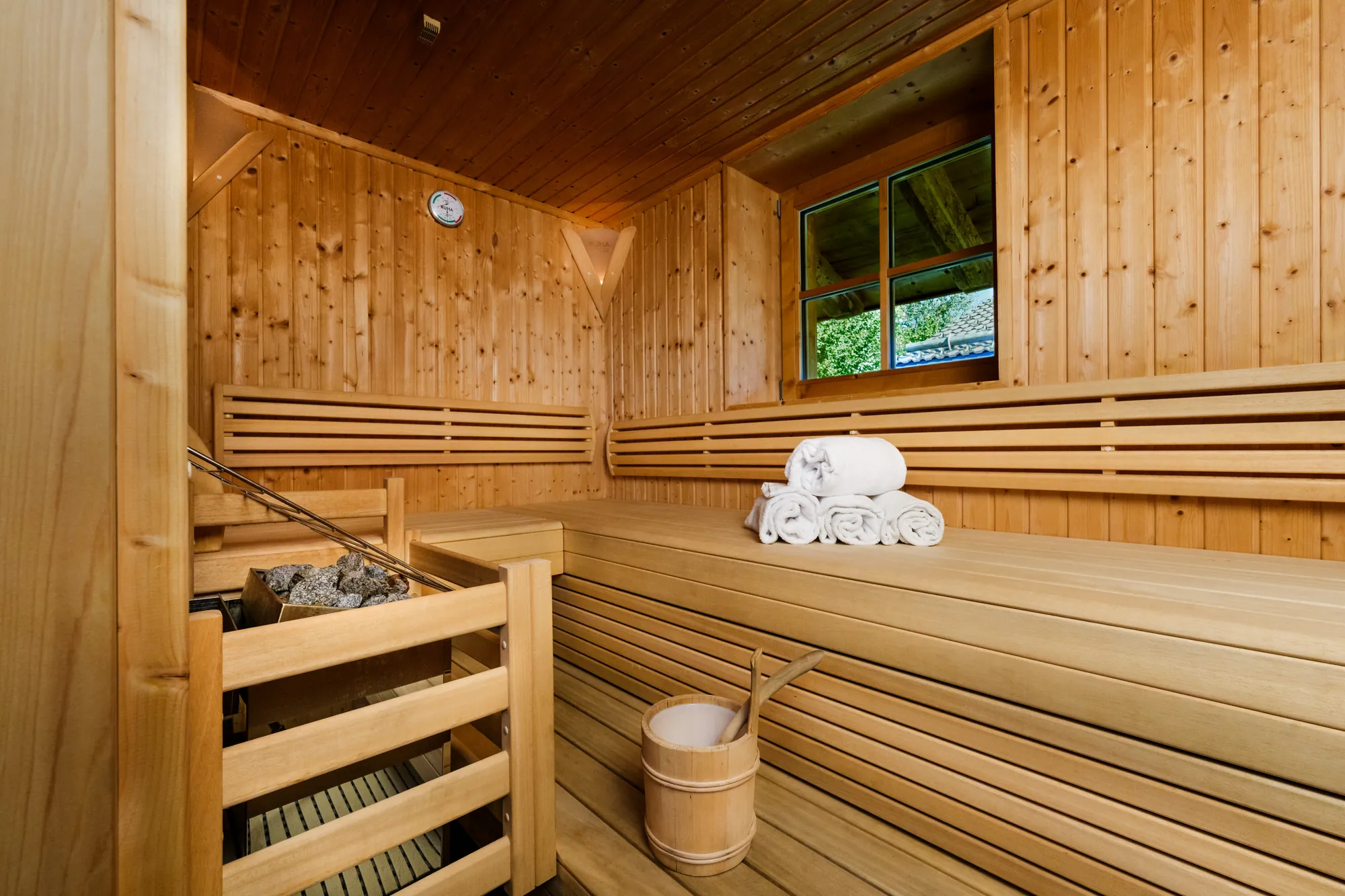 Finnische Sauna aus hellem Holz mit Liegebänken, Saunaofen, Handtüchern, Kübel und kleinem Fenster