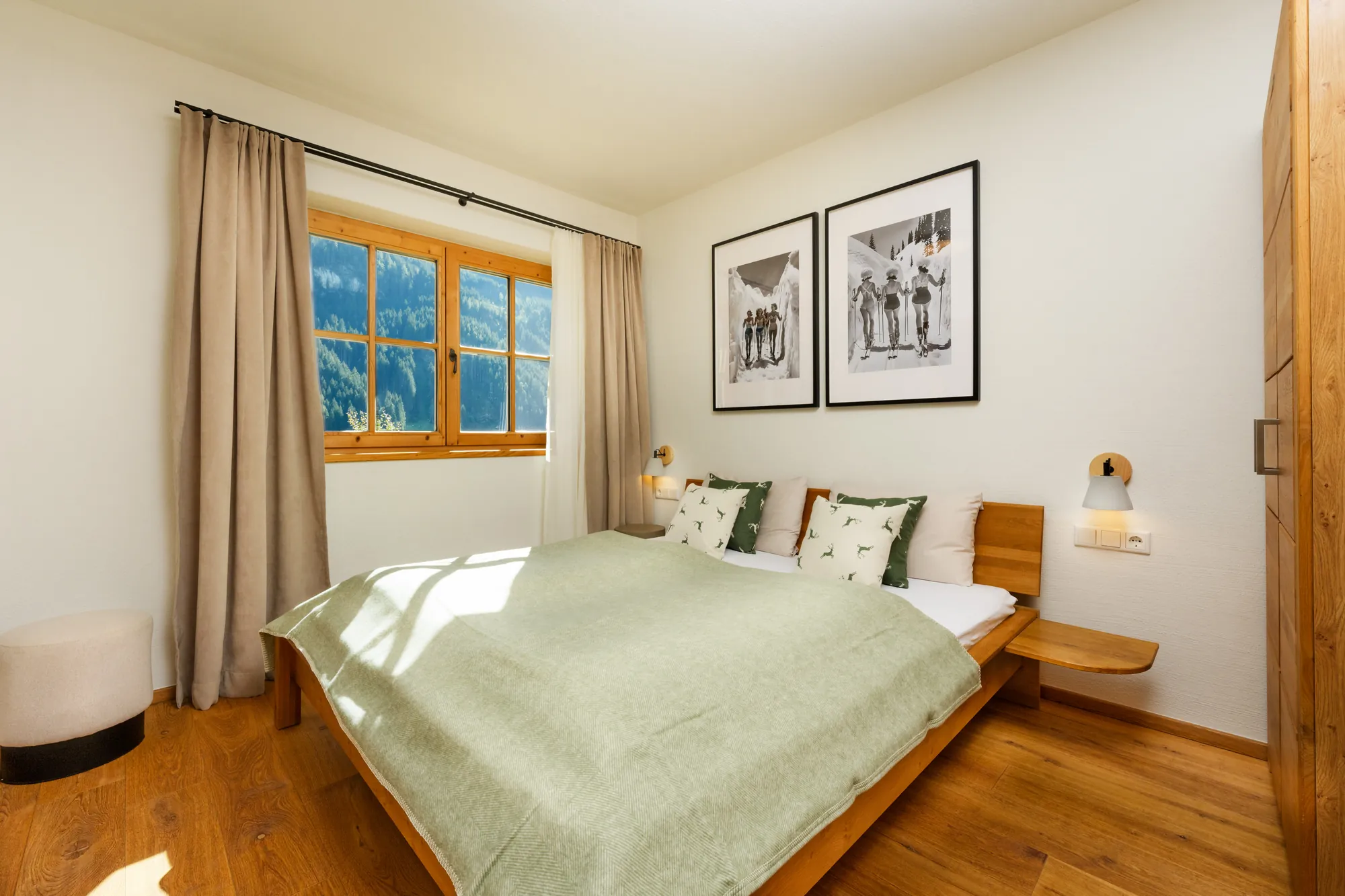 Schlafzimmer mit Kingsize-Bett aus Holz, grüner Tagesdecke, Hirschkissen, Schwarz-Weiß-Skifotos und Fenster mit Bergblick