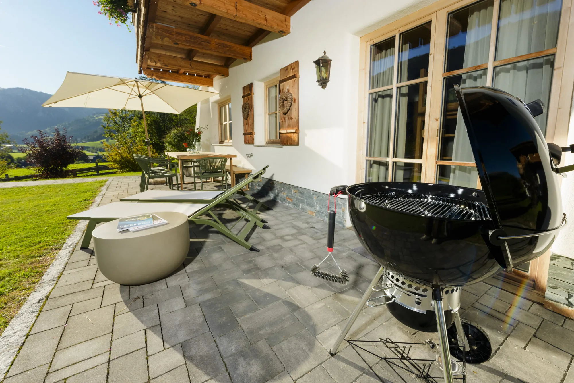 Terrasse des Chalet Gams mit schwarzem Kugelgrill, Sonnenliegen, Sonnenschirm und Blick in den Garten