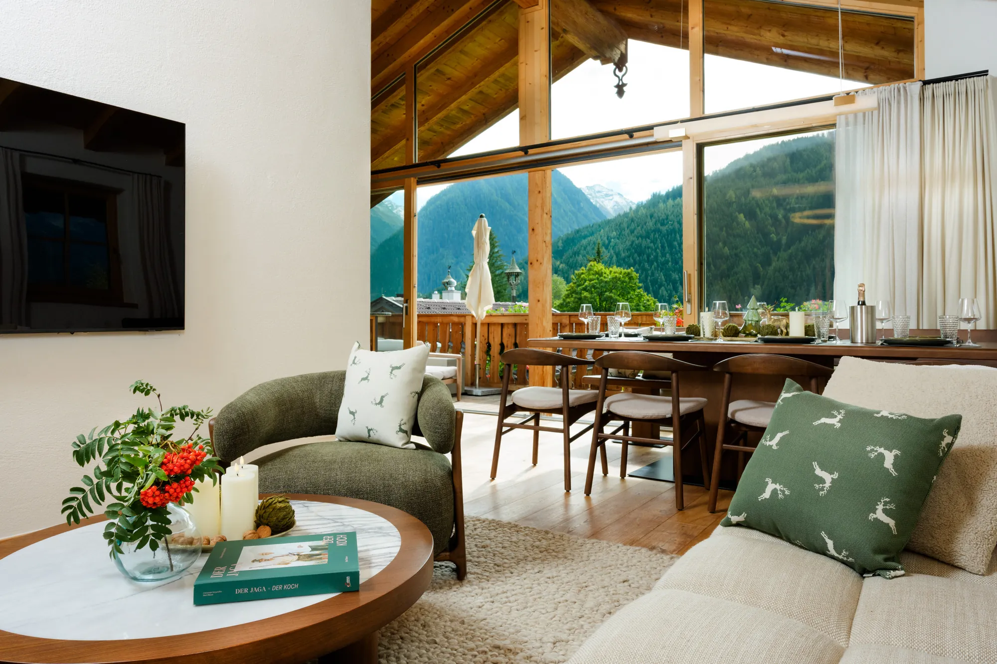 Sessel und Couchtisch im Wohnbereich, dahinter Esstisch und große Glasfront mit Blick auf Balkon und Berge